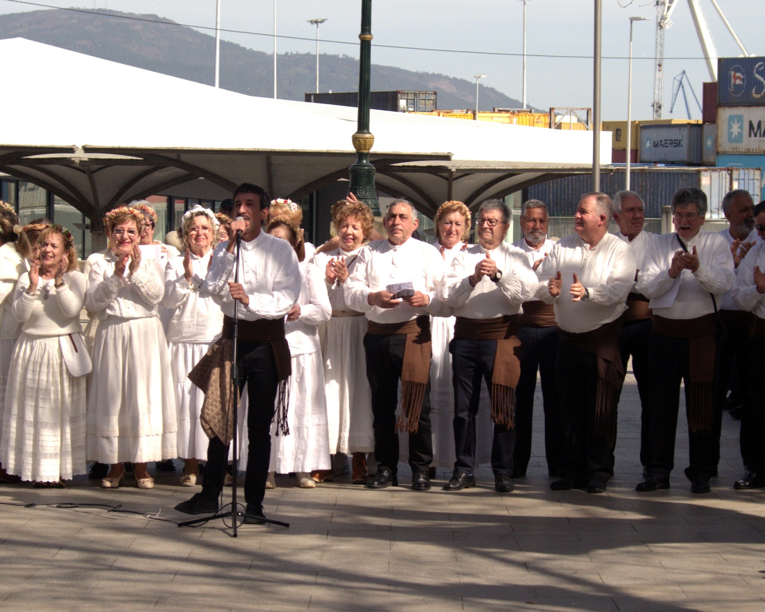 Grupo de cantareiras e cantores interpretando música tradicional galega nun escenario exterior.